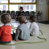 Quatre jeunes enfants sont assis par terre face à un adulte à l'intérieur d'une salle de classe, avec des cordes vertes pour la danse contemporaine école disposées sur le sol et des chaises vides alignées contre le mur.