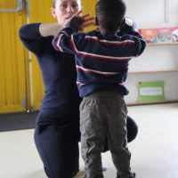 Dans une salle de classe aux murs et aux étagères jaunes, un adulte est agenouillé face à un enfant qui se tient devant lui ; tous deux lèvent les mains près de leur visage comme s'ils pratiquaient des mouvements inspirés de la danse contemporaine école.