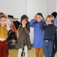 Un groupe de jeunes enfants se tient dans une salle de classe, fabriquant des lunettes avec leurs mains sur leurs yeux tout en participant à une activité inspirée de la danse contemporaine école.